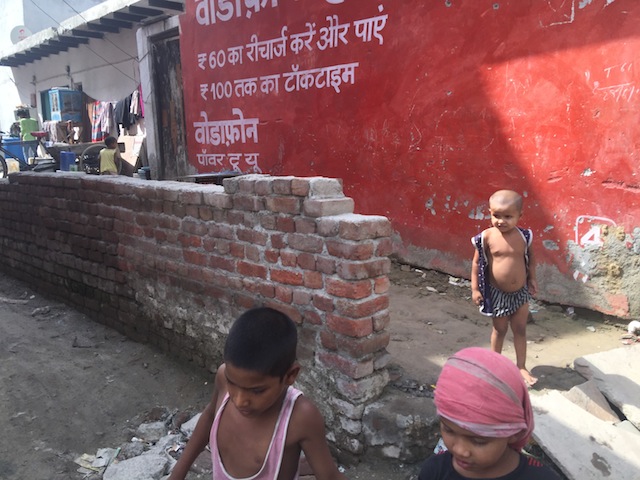 Malnourished kids run around outside a ration shop in India. The lettering on the side of the building is part of an advertisement by a multinational telecom company, peddling cheap phones in the country that hosts the world’s largest population of hungry people. (Photo: Neeta Lal / IPS)