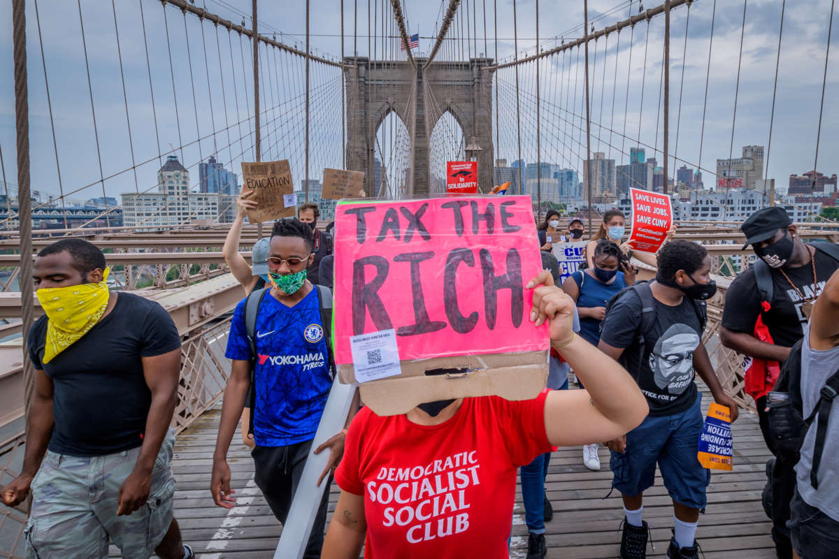 A protester in a red DEMOCRATIC SOCIALIST CLUB t-shirt holds a sign reading "TAX THE RICH" at a protesy\t