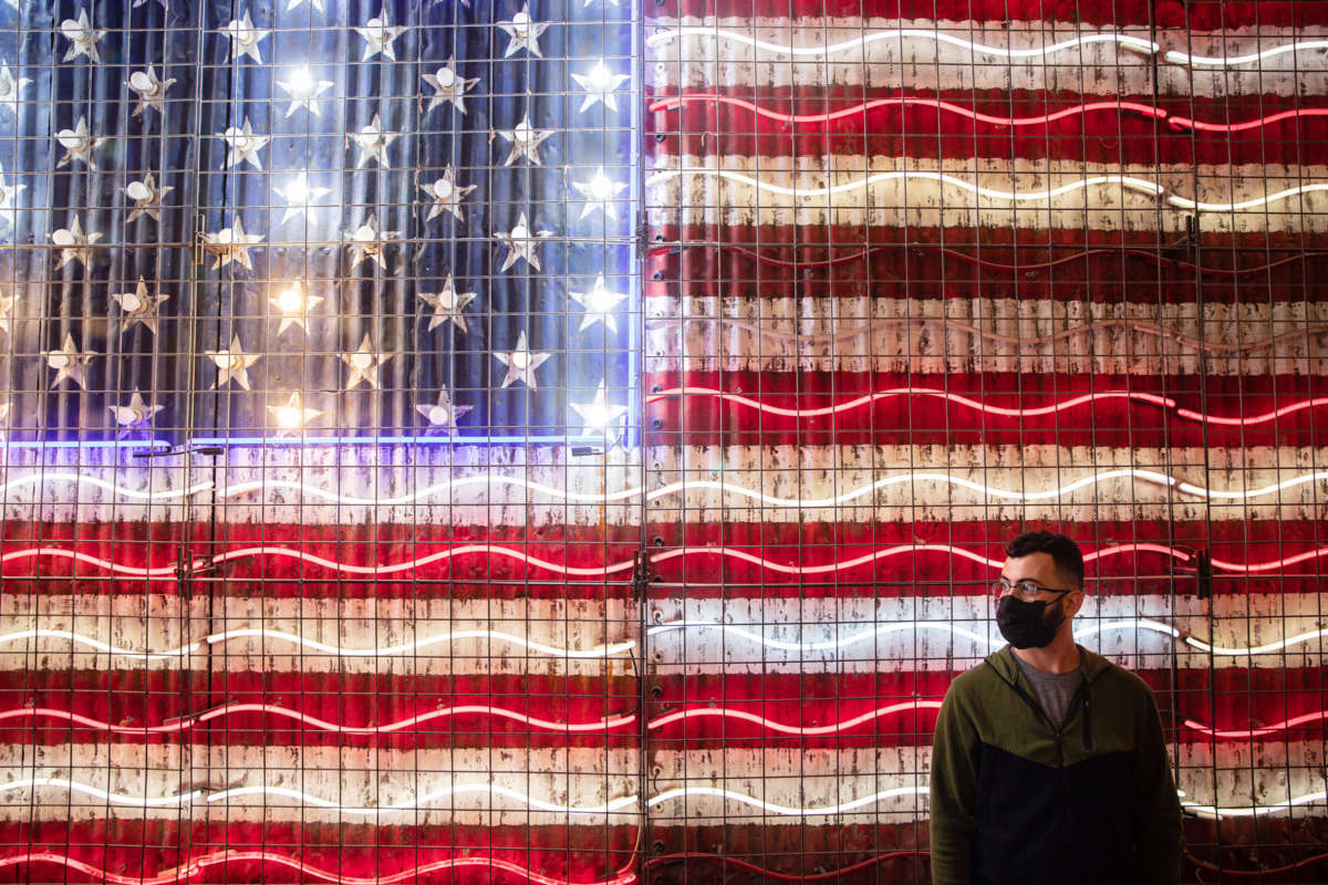 A masked man waits in front of a neon sign resempling the U.S. flag