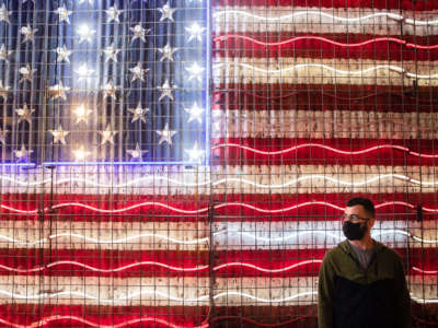 A masked man waits in front of a neon sign resempling the U.S. flag