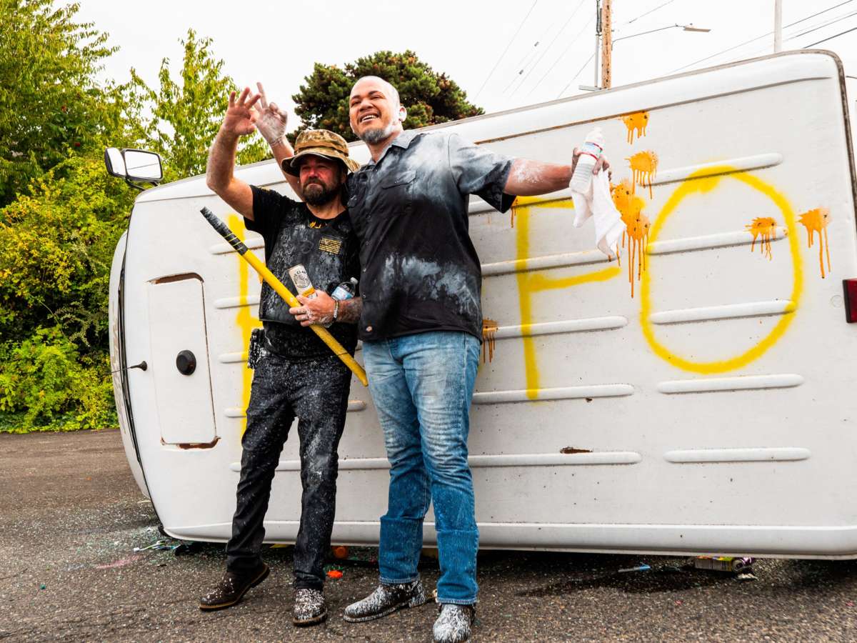 Two pant-smeared men pose in front of a tipped-over van