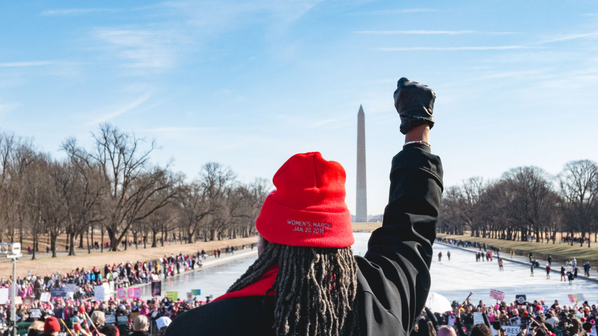 A woman stands on the steps of the Lincoln Memorial, facing the crowd and the Washington Monument, holding her fist up, and wearing a knitted cap with "Women's March Jan. 20, 2018" imprinted on it.