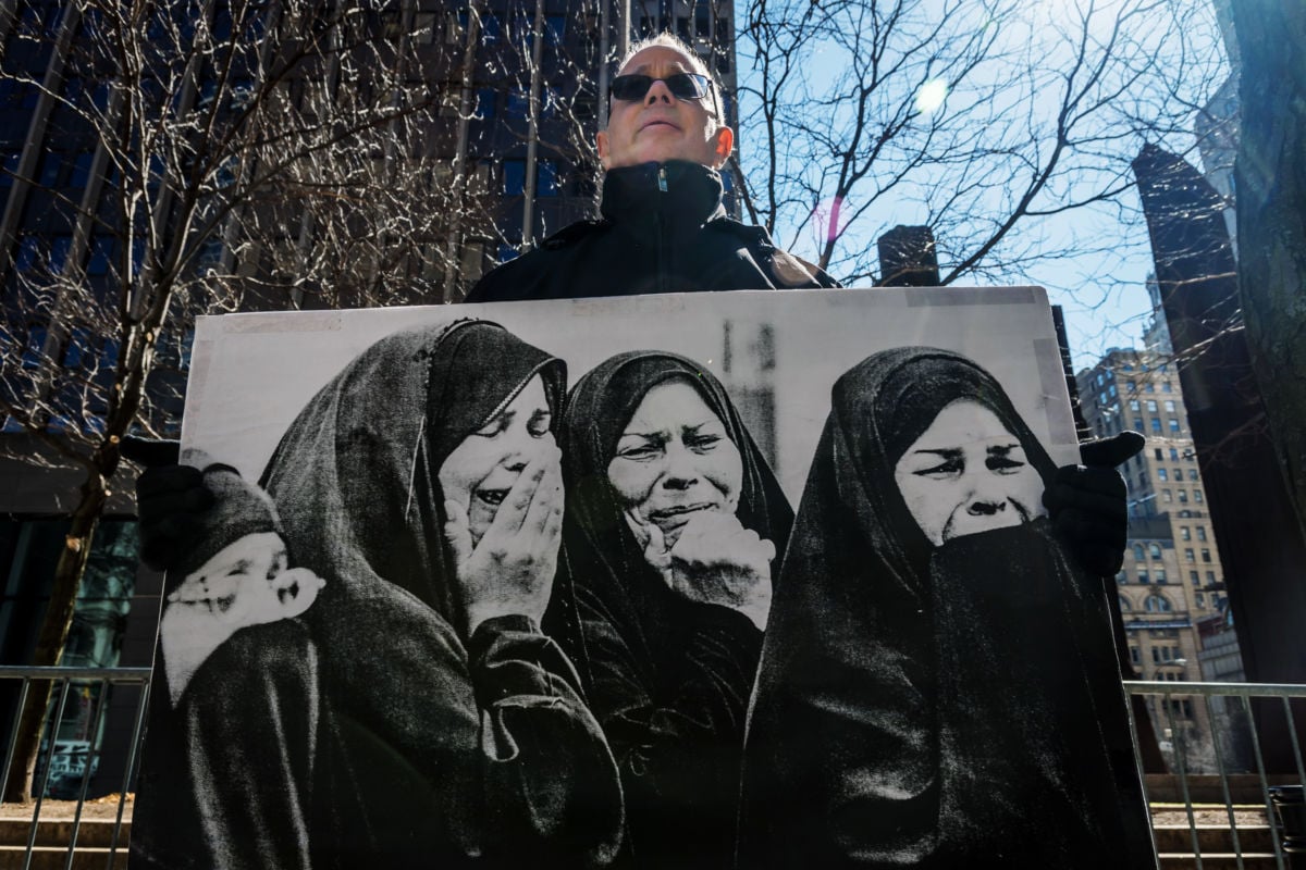 An older man holds a black and white photo of crying women in chador