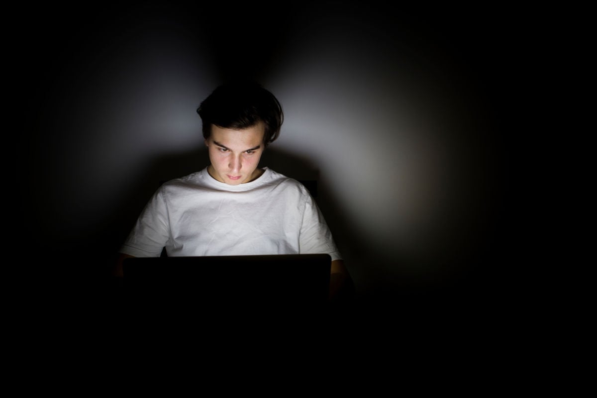 A young white man is illuminated by his laptop's screen in a darkened room