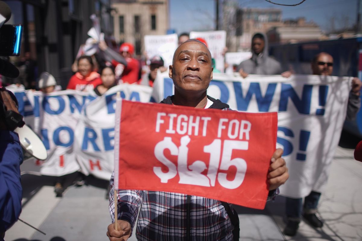 A man stands ahead of a protest holding a red sign reading "FIGHT FOR $15"