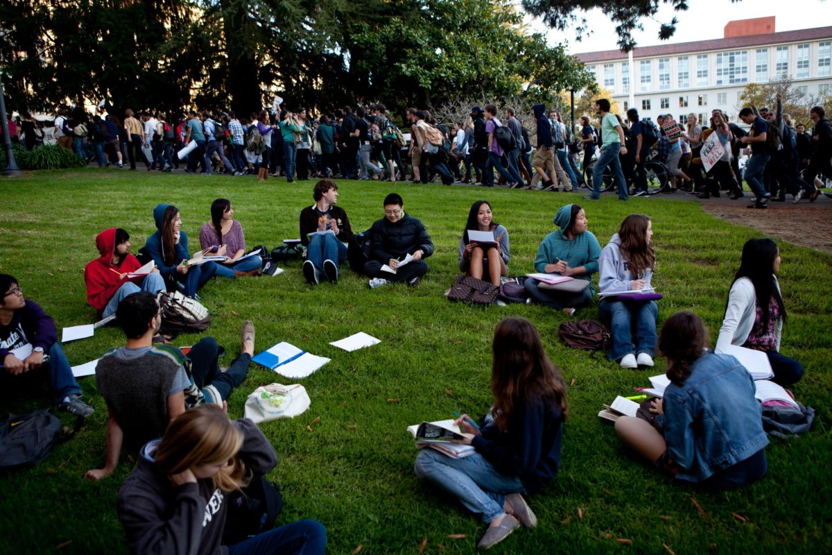 Students sit in a circle on a grassy lawn as people walk to a protest behind them