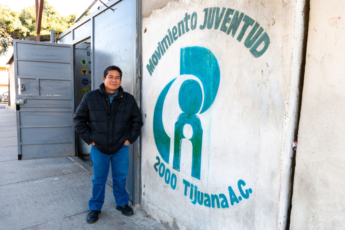 A man in a black jacket stands next to a wall reading "MOVIMENTO JUVENTUD 2000 Tijuana A.C"