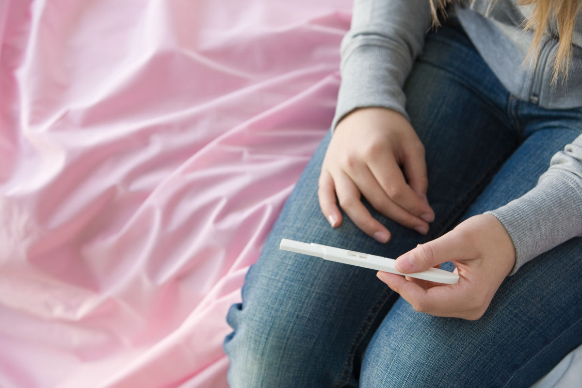 A girl sits on a bed and looks down at a pregnancy test