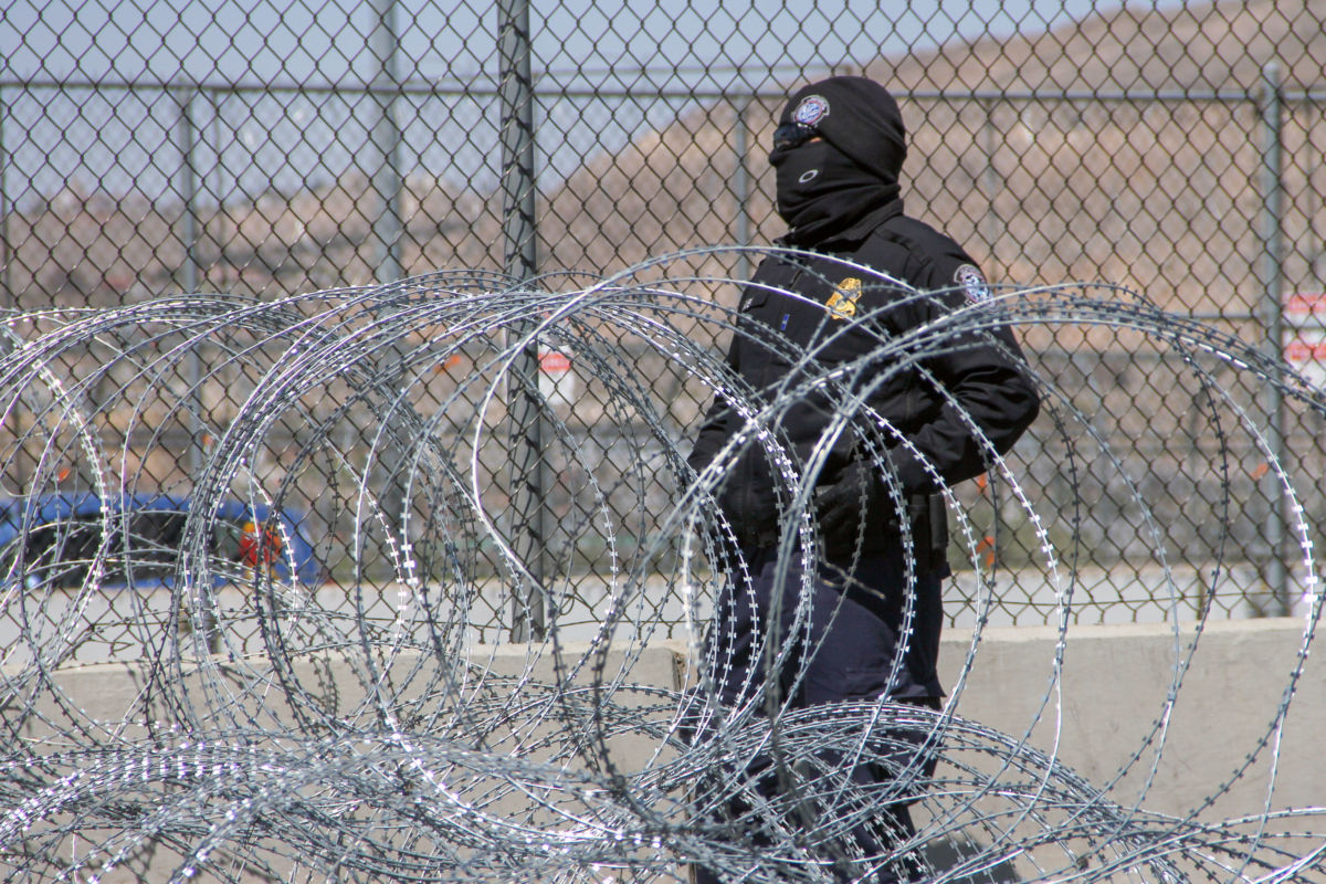 A Border Patrol guard wearing all black stands surrounded by razor wire