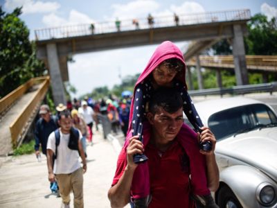 A father carries his daugher on his shoulders as he walks with other migrants down a highway