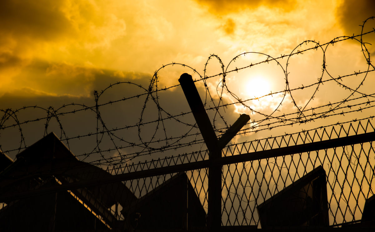 A barbed wire fence is seen backlit from a yellow sunset, prison