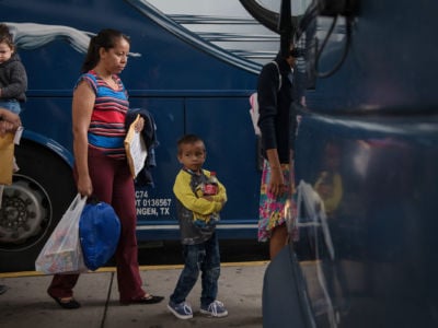A young boy walks with his mother toward a greyhound bus, migrant detention