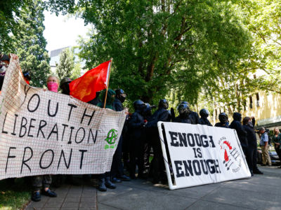 People clad in black stand behind signs reading ENOUGH IS ENOUGH and YOUTH LIBERATION FRONT