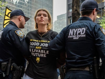 A protester wearing a shirt reading "GOOD JOBS AND A LIVABLE FUTURE" is carried away by cops