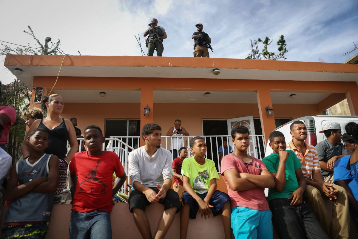 A row of Puerto Rican people sit on a retaining wall as soldiers carrying guns overlook them from a rooftop behind them