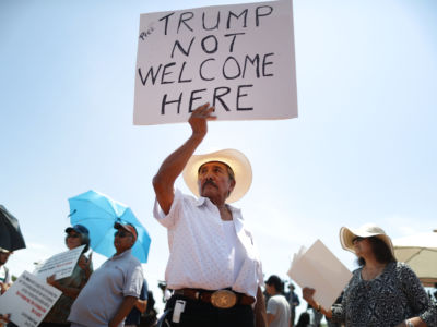 A man in a cowboy hat holds a sign reading "PRES. TRUMP NOT WELCOME HERE" during a protest