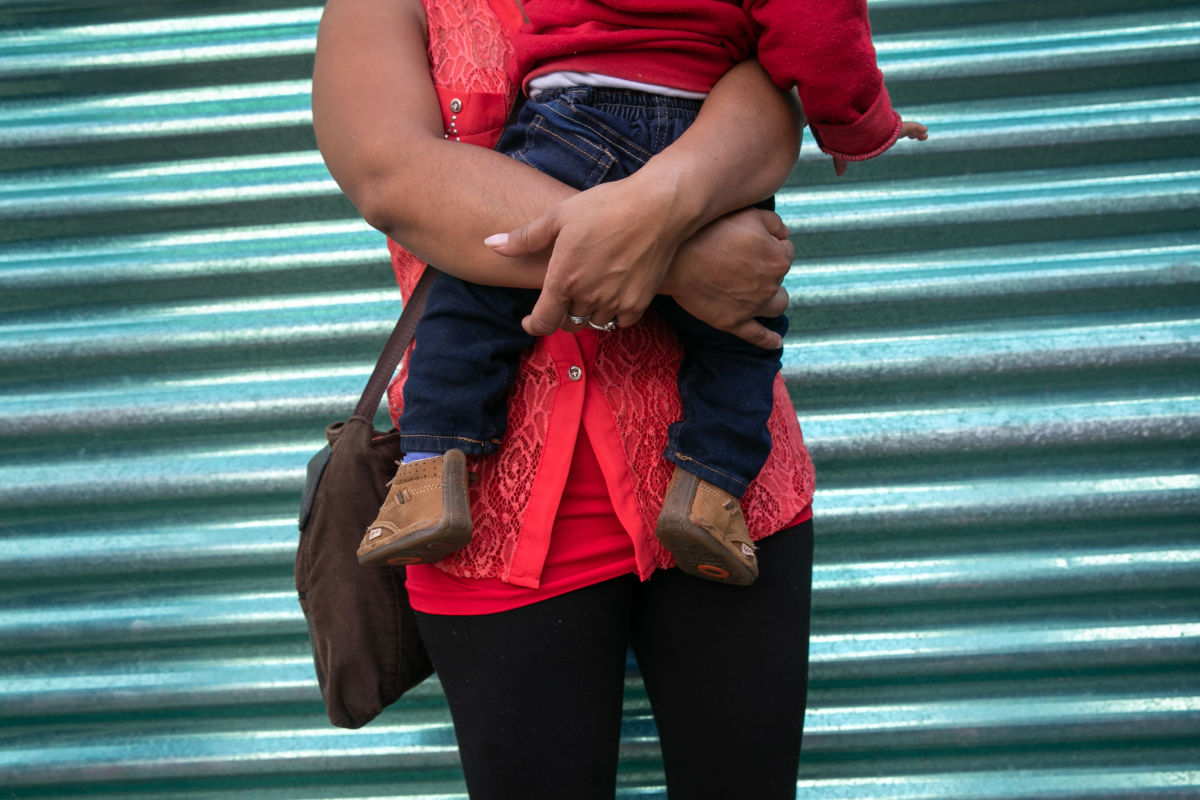 A mother holds her child while standing in front of a corrugated metal fence