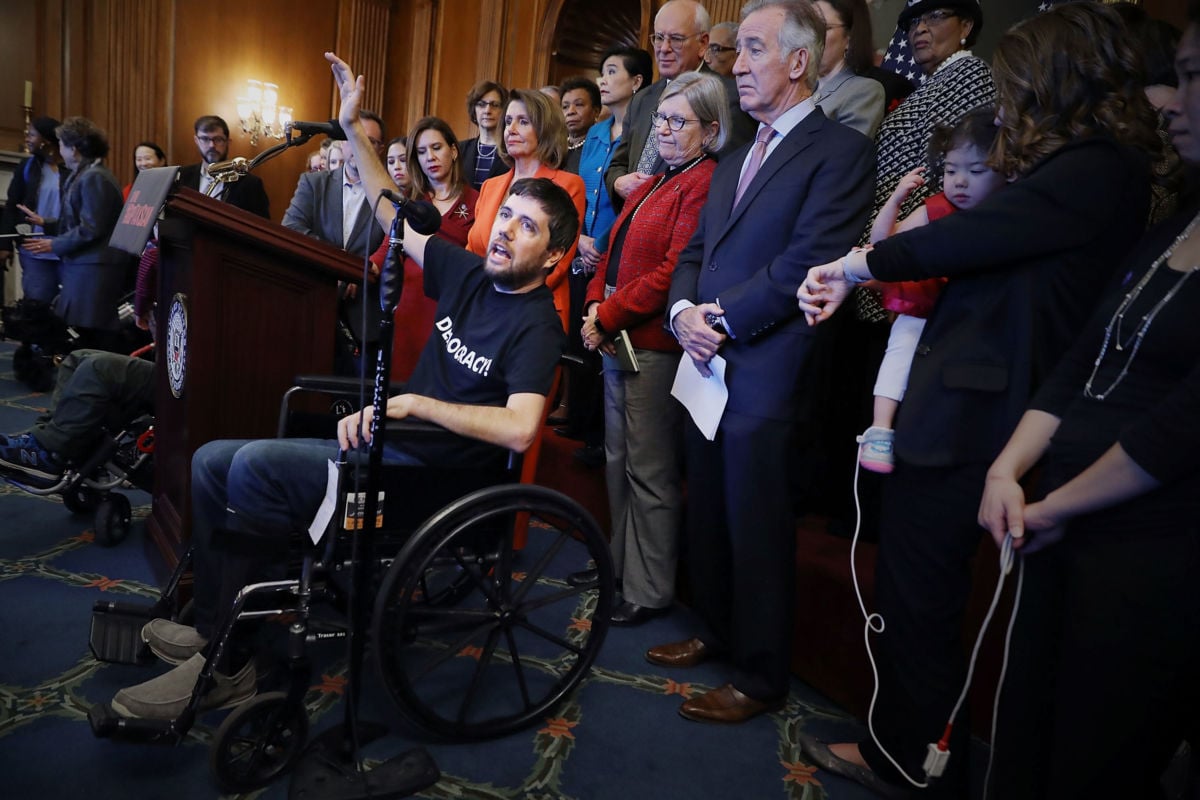 A man wearing a shirt reading "Democracy!" speaks into a microphone while seated in a wheelchair