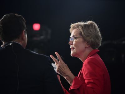 Elizabeth Warren speaks to members of the press in a darkened room, debate