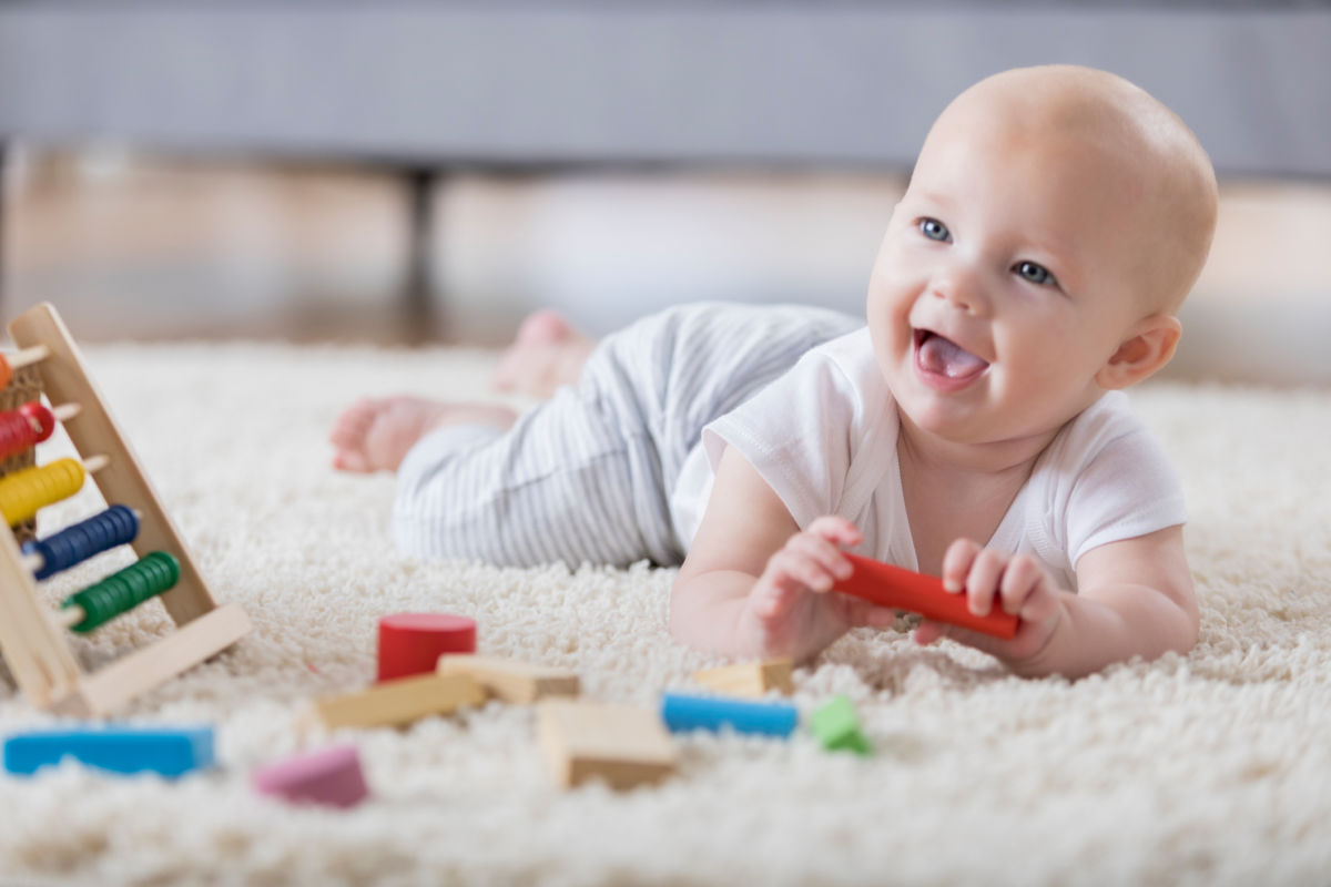 A cute baby lays on her tummy on a rug in a living room and sings with an open mouthed smile. She is holding one of several wooden blocks. There is an abacus in the foreground.
