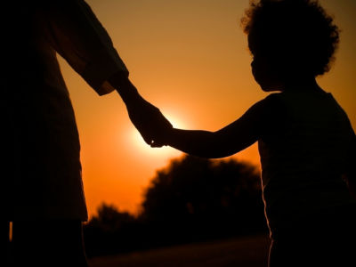 A Black child holds an adult's hands while silhouetted by the sunset