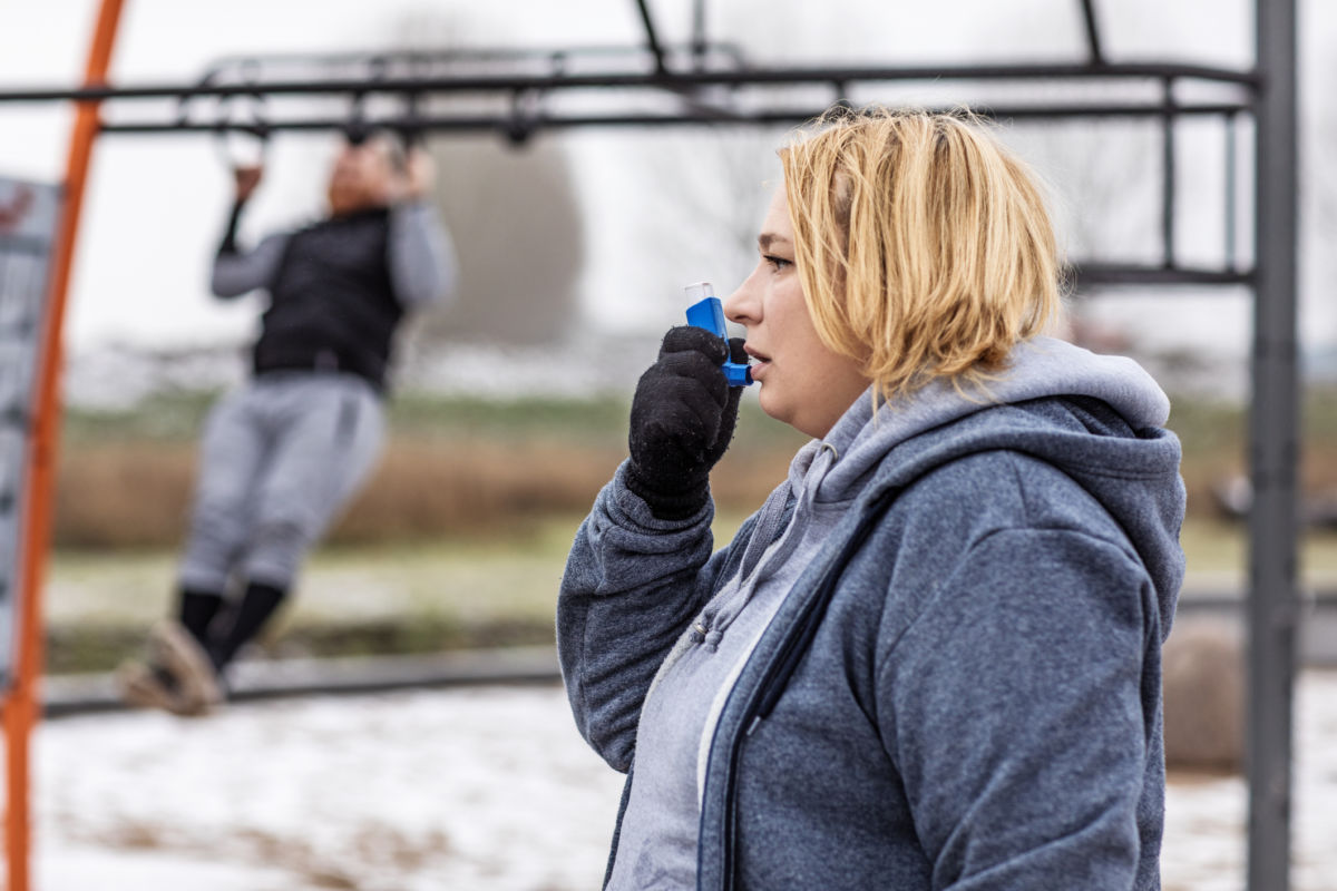A blonde woman takes a puff of her inhaler while someone does pullups on the structure behind her