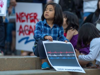A young girl sits on the sidewalk during a protest while holding sign depicting red, white and blue lengths of barbed wire