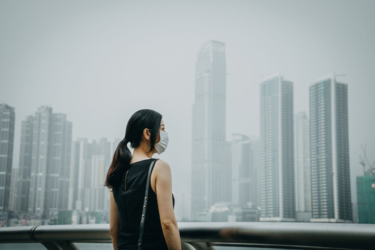 A young woman with a face mask on stands in front of an extremely smoggy city