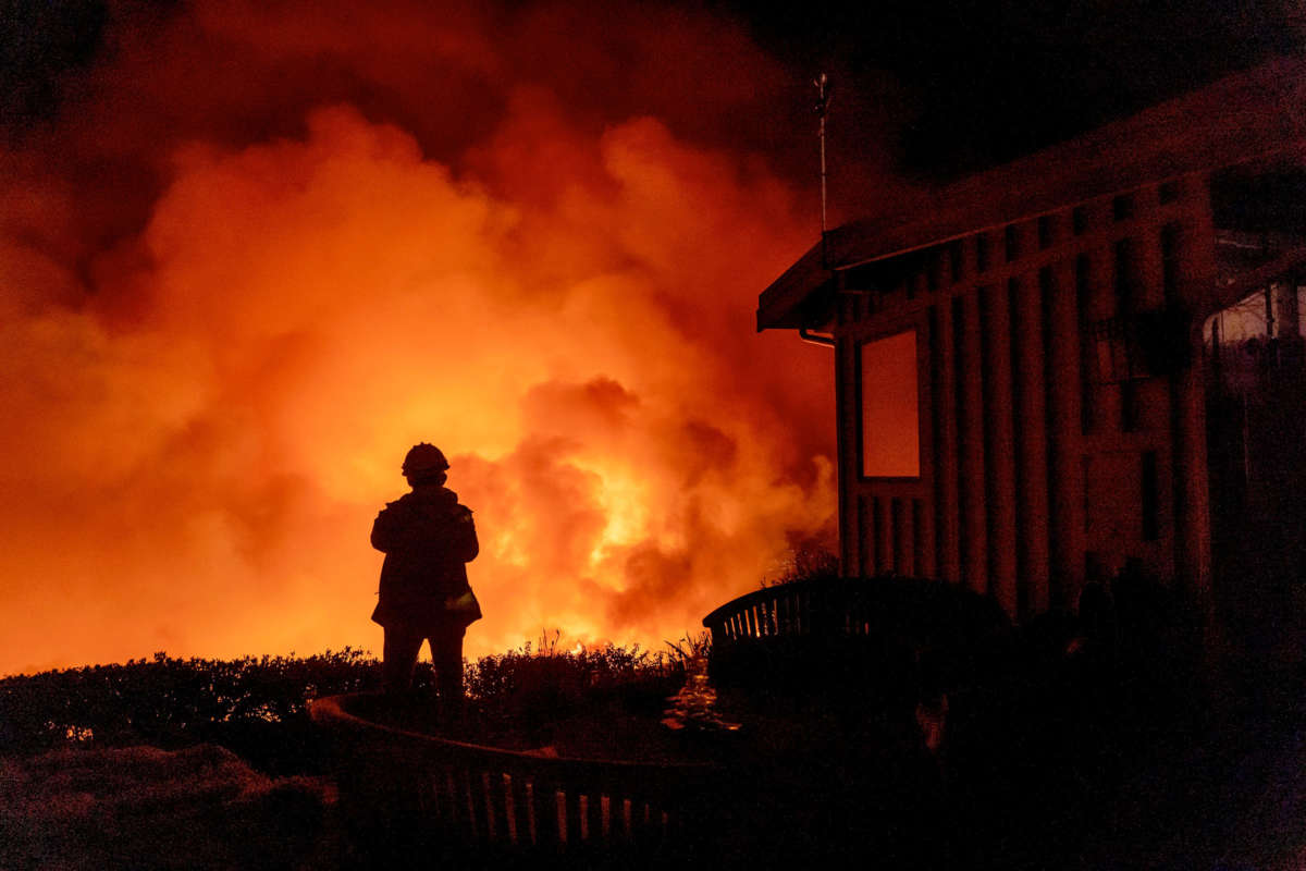 A person stands by a house and watchs a wildfire blaze