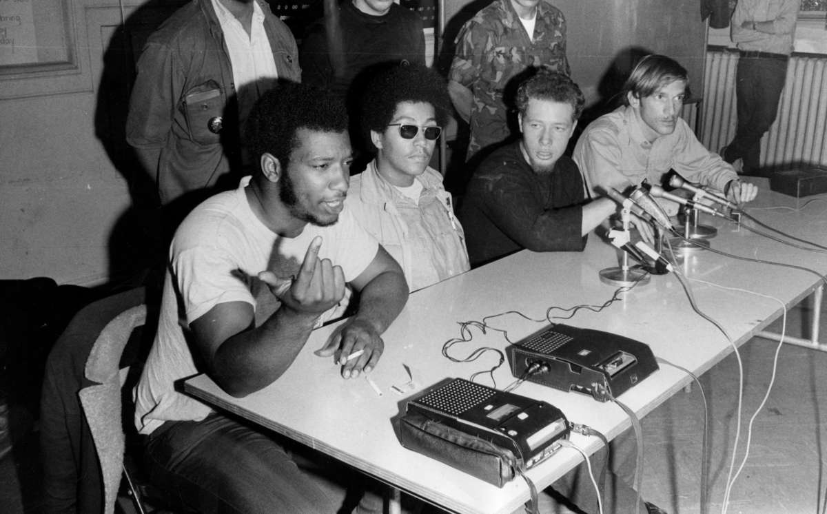 Activist Fred Hampton speaks into a microphone while seated at a table with others