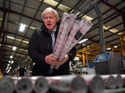 Boris Johnson stands in a factory holding rolls of wrapping paper