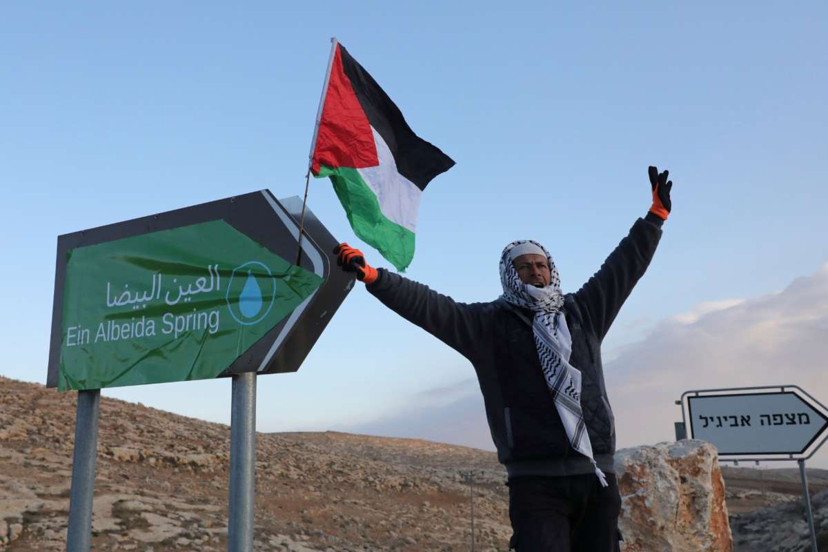 A man raises his arms in triumph next to a sign reading "Ein Albeida spring"