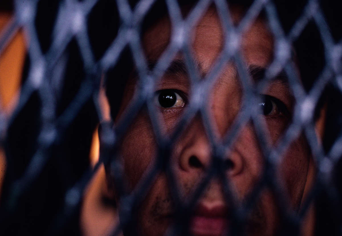 A man looks out from the spaces in the mesh fence separating him from the camera