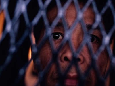 A man looks out from the spaces in the mesh fence separating him from the camera