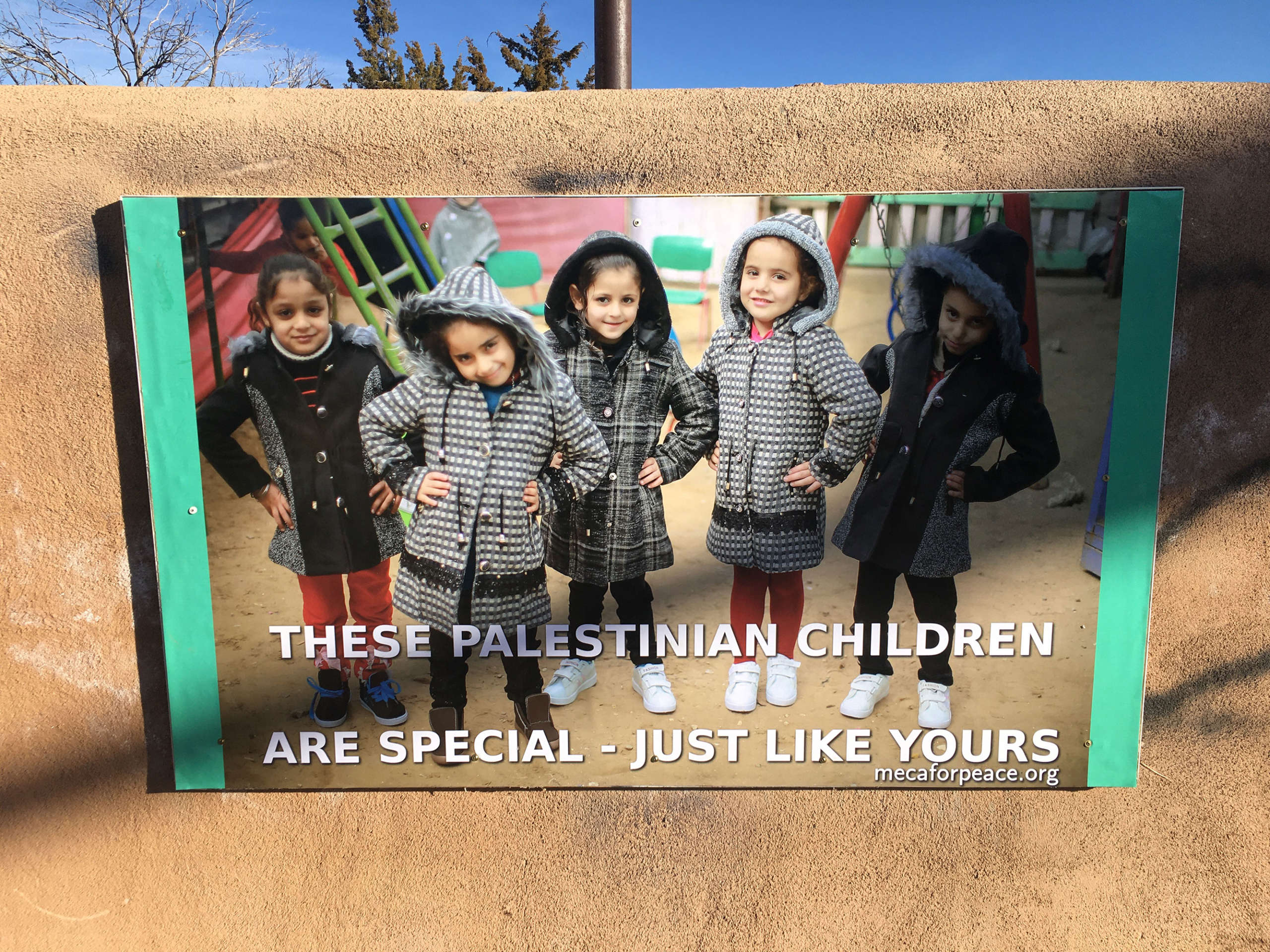 A sign displays five 6-year-old Palestinian girls in winter coats with the caption, These Palestinian Children Are Just Like Yours.