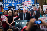 Bernie sanders speaks into a microphone surrounded by people holding "Medicare for All" posters