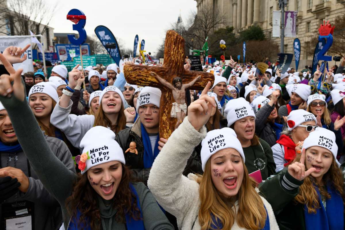 white women in white hats march down the street carrying a cross