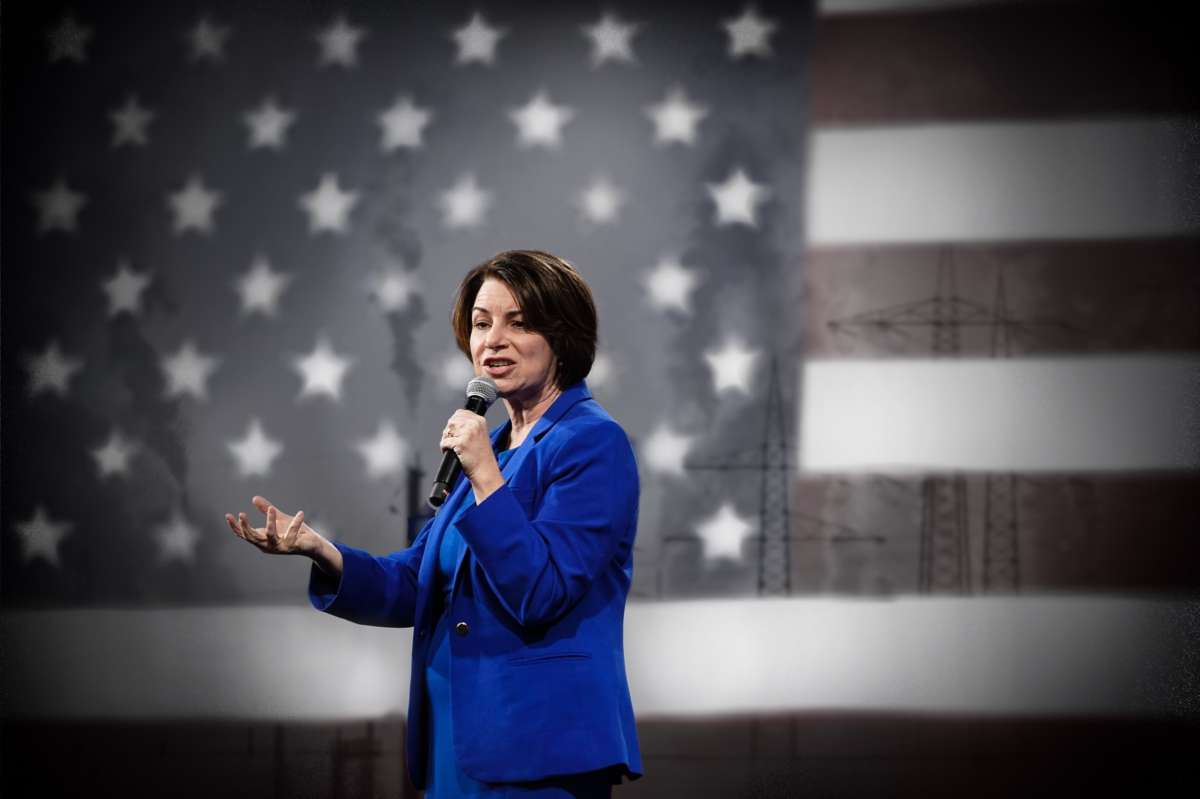 Amy Klobuchar speaks into a microphone in front of a U.S. flag that's superimposed with smokestacks in the distance