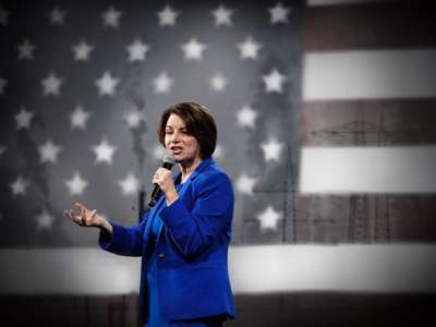 Amy Klobuchar speaks into a microphone in front of a U.S. flag that's superimposed with smokestacks in the distance