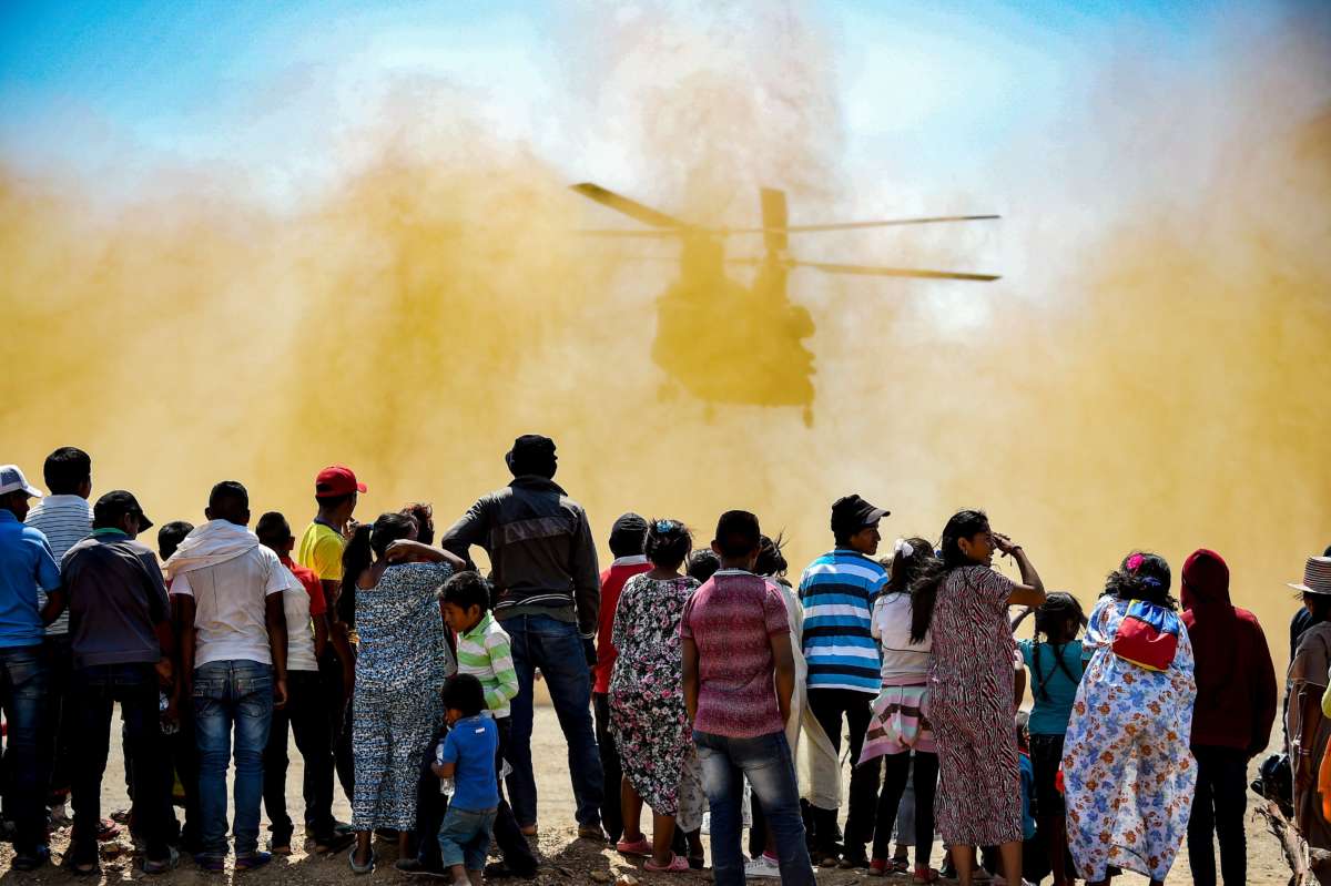 A line of people stand and watch as a us army helicopter kicks up dust upon its departure
