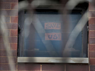 A sign reading "SAVE US" is displayed in a prison window festooned with razor wire