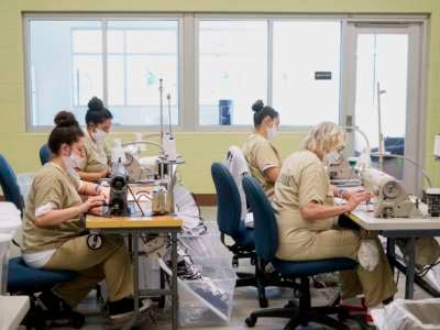 Four women in two rows construct face masks at workstations reminiscent of a sweatshop