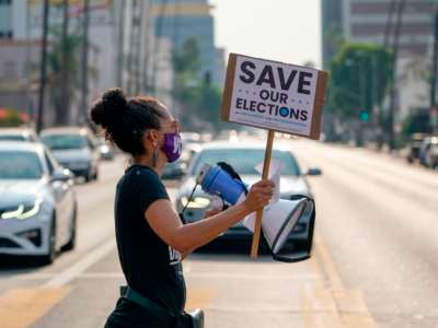 A protester crosses a street holding a sign reading "SAVE OUR ELECTIONS"