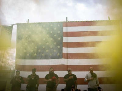 White men hold their maga hats over their hearts as they stand in front of a us flag, yellow smoke billowing in the foreground