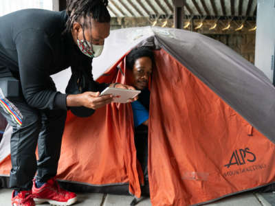 A person in a mask stoops down to help a homeless man in a tent fill out the census on a tablet
