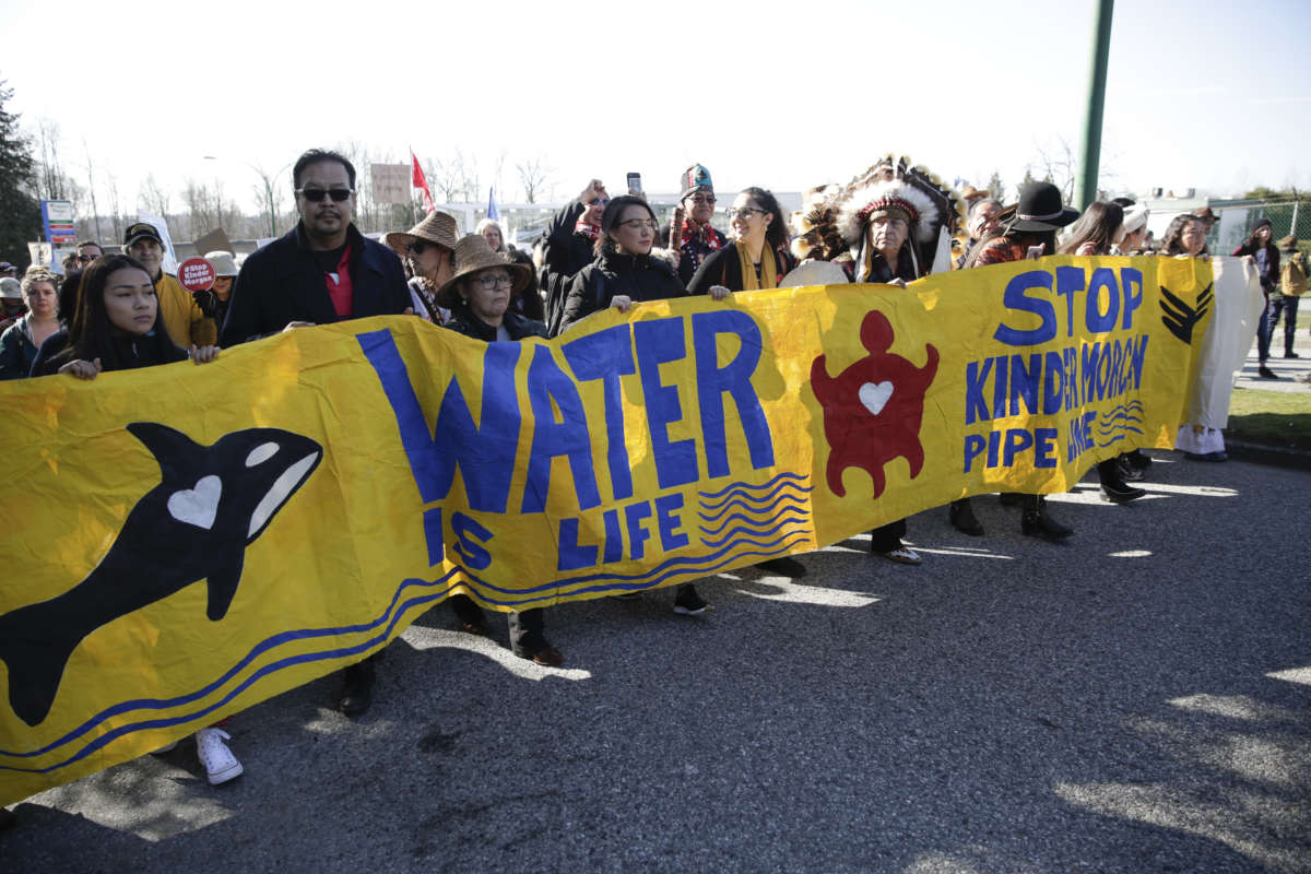 People march behind a banner reading "WATER IS LIFE, STOP KINDER MORGAN PIPELINE" during an outdoor pre-covid protest