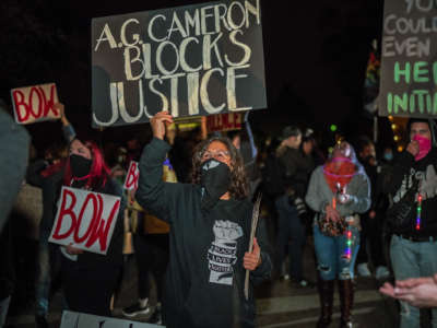 A protester holds up a sign reading "AG CAMERON BLOCKS JUSTICE" during a protest