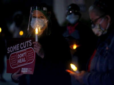 Nurses hold a candle light vigil, one holding a sign reading "SOME CUTS DON'T HEAL"