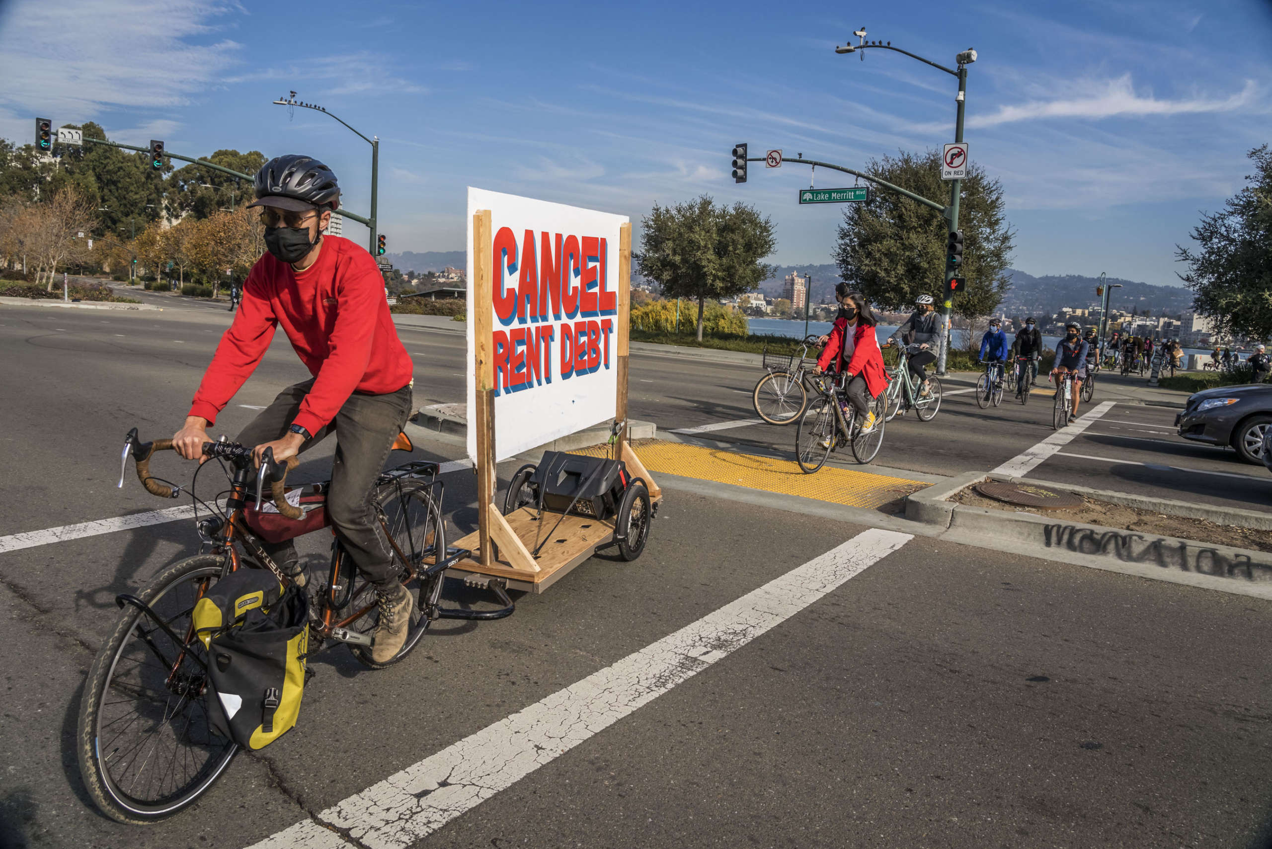 En karavane med sykler og biler forlater Lake Merritt, mens hovedsykkelen trekker et banner med handlingens krav: Cancel the Rent.