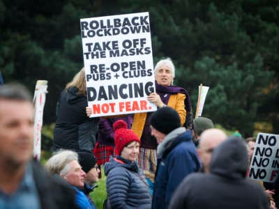An unmasked protester holds a sign reading "ROLLBACK LOCKDOWN, TAKE OFF THE MASKS, RE-OPEN PUBS + CLUBS, DANCING NOT FEAR." during a protest
